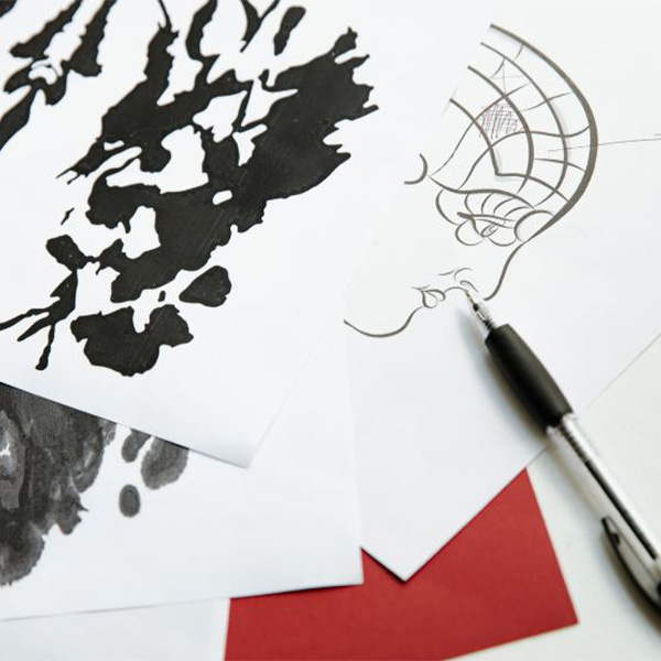 A pen laying next to papers with images of a side profile drawing and a rorschach inkblot test.
