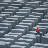 Photograph of the Holocaust Memorial in Berlin: a landscape of hundreds of large, jagged gray cubes, with a person in a bright red jacket stepping across a gap