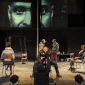 Photograph of a stage, with a camera pointed at an actor whose face is projected on the back wall