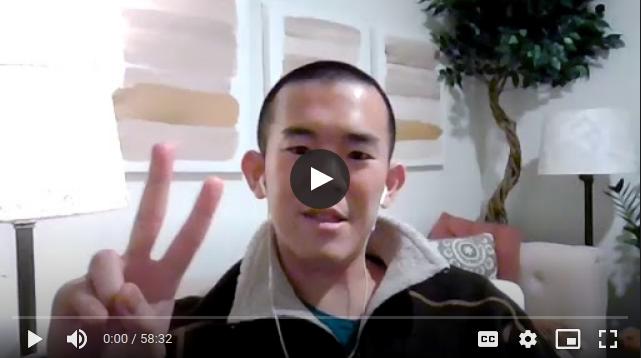 Still frame from a video: man speaking into the camera while holding up two fingers of his right hand. Indoors, with a small ornamental tree in the background.