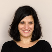 Woman with black hair and a black shirt, smiling to the camera against a bright beige background