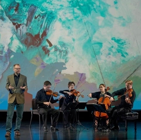 String quartet and male speaker, seated and standing respectively in front of a large projection of an abstract painting in blue-green hues