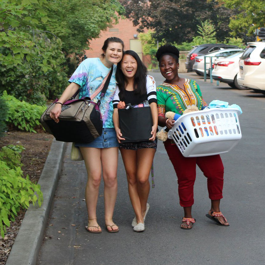 Three students smile at the camera while moving their belongings into their residence hall.