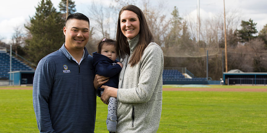 Brian and Kirsten Kitamura hold their baby while standing in a baseball field.