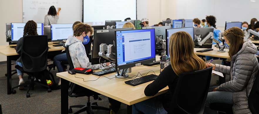 Image description: photograph of students sitting in a computer lab with a professor writing on a whiteboard in the background