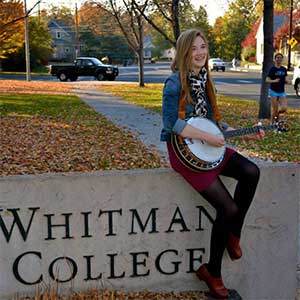 A student sits on Whitman College sign playing a banjo with fall colors in the background
