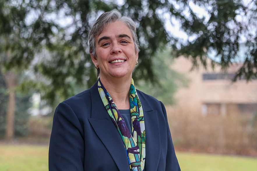 Image description: photo of Dr. Sara Bolton, a smiling woman with short grey hair wearing a navy blue jacket and green, purple and blue scarf against a blurred background of trees and buildings