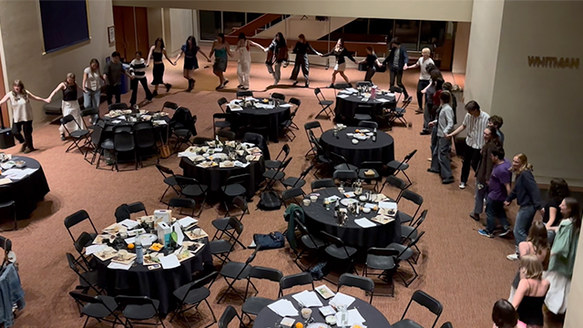 A large circle of people holding hands surrounding several round tables with black tablecloths in Cordiner Hall lobby..