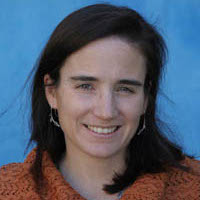 Woman with light skin and dark hair, smiling against a blue backdrop.