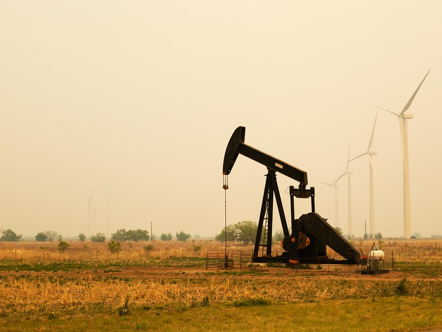 Photograph of an oil derrick, with windmills in the background against a smoky sky