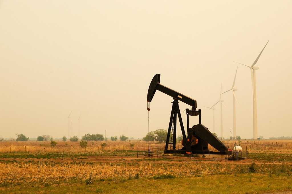 Photograph of an oil derrick, with windmills in the background against a smoky sky