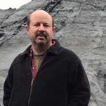 Middle aged white man in light outdoor clothing, standing on a glacier under a bright gray sky