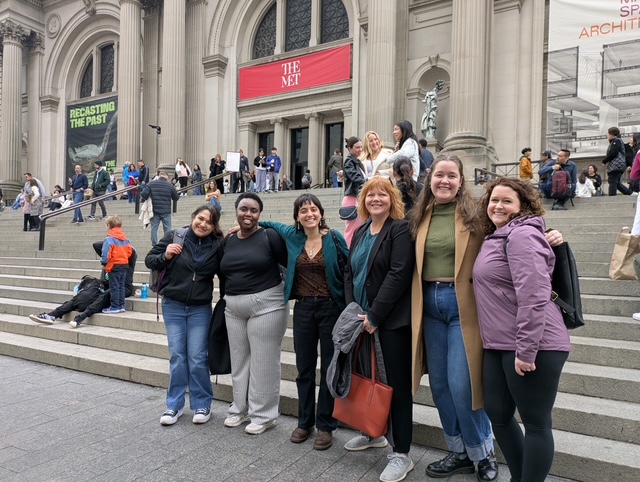 A group of people stand together in front of the steps at The Met in New York City. They are wearing coats and winter clothes and smiling at the camera.