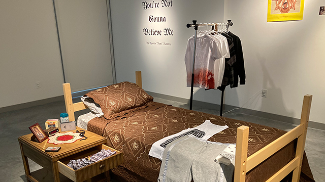 A bedroom scene set up in a gallery space with the words “You’re Not Gonna Believe Me" on the wall behind.