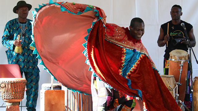 A dancer in the center twirls a bright red cape while two performers in the background play percussion instruments.