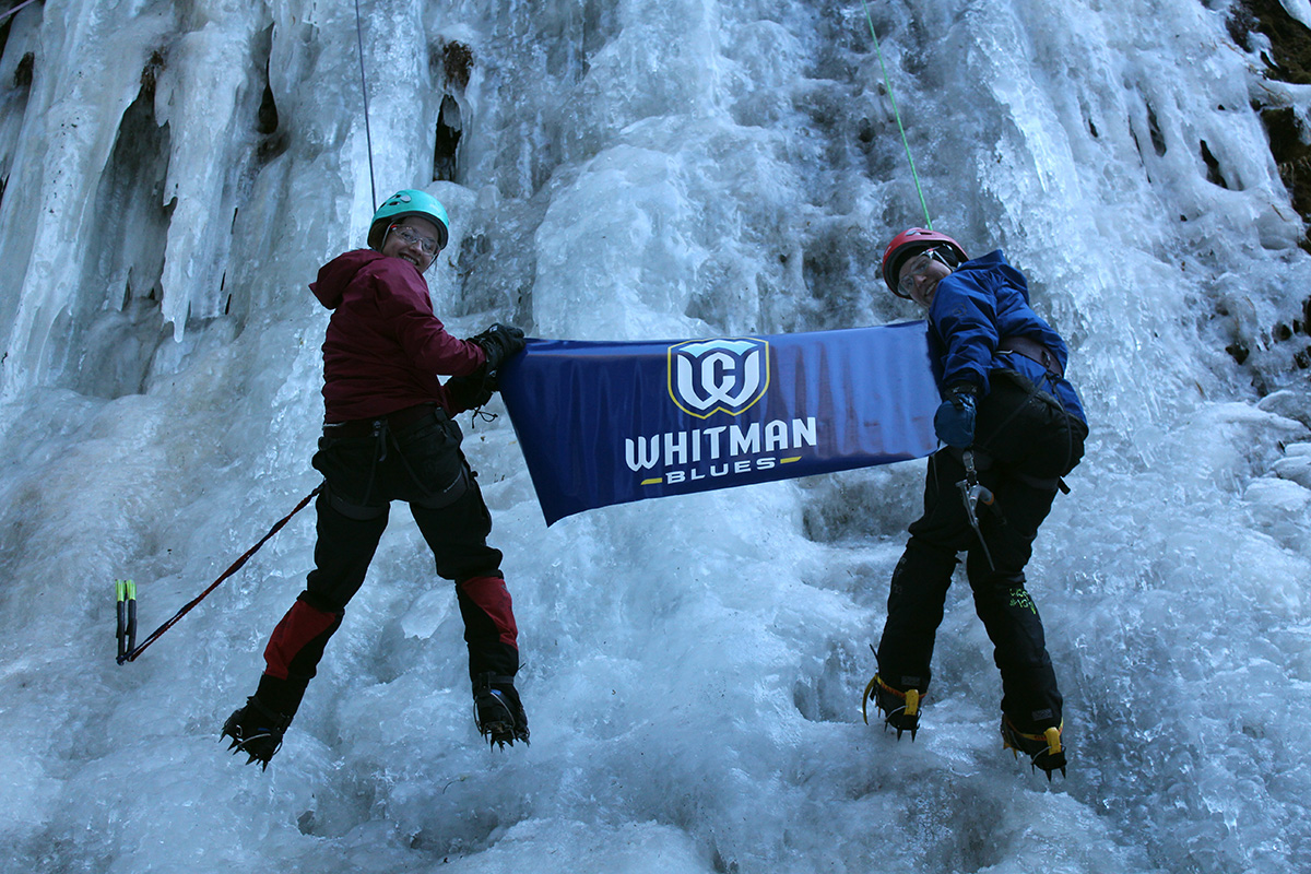 Two Whitman students holding up a Whitman Blues banner while ice climbing.