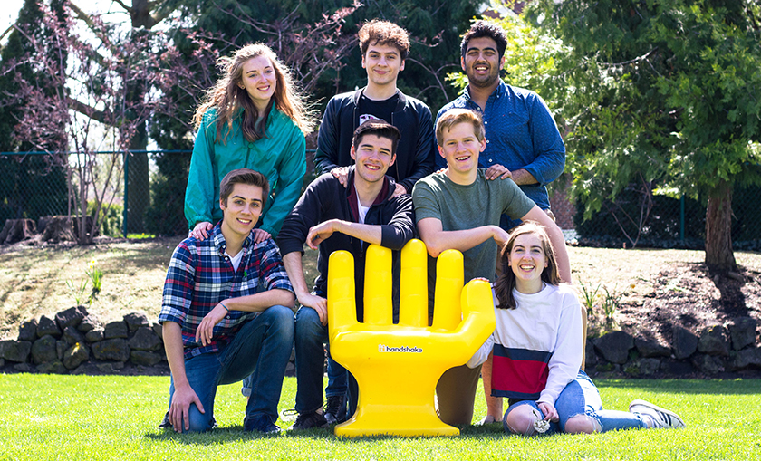 Seven students pose with the yellow Handshake chair on the Reid side lawn by College Creek.  