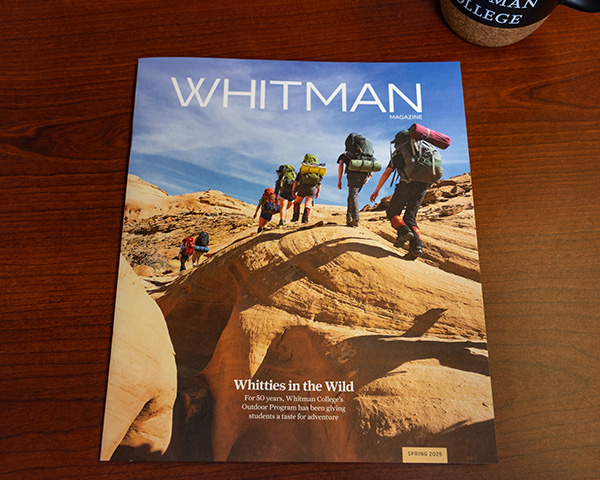 A magazine with a cover of students hiking sits on a wood table with a Whitman College mug nearby