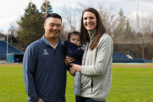 Brian and Kirsten Kitamura hold their baby while standing in a baseball field.