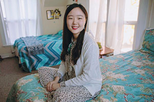 Esther Ra smiles while she sits on a bed.