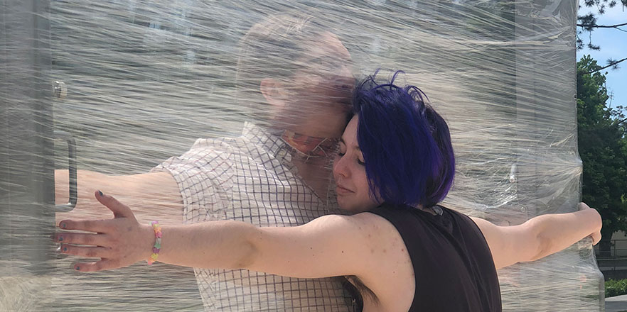 Two people hug with a sheet of plastic wrap between them.