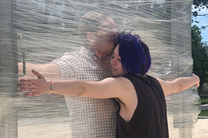 Two people hug with a wall of cellophane between them.