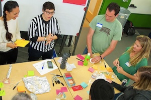 Katie Krummeck wears a black and white striped referee shirt as she leads a group of educators in an activity involving many colored post-it notes, pens and other items 