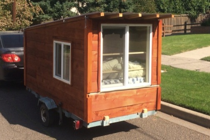 a tiny house with white windows is pulled down a road behind a car