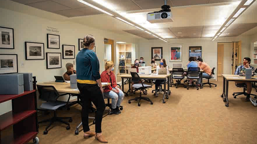 Photo of the new Edwards Classroom in Whitman College's Penrose library. Several students are seated at desks facing the camera. A professor stands at the head of the classroom with her back to the camera.