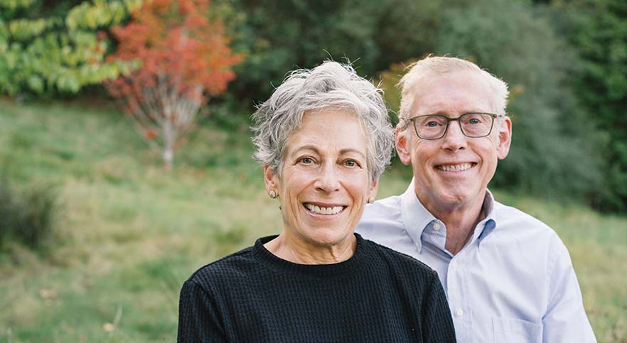 Photo of Nancy and Greg Serrurier, seated outside with trees bearing fall colors behind them.