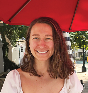 Alex Feller with shoulder-length brown hair and wearing a white top, smiles under a red canopy.