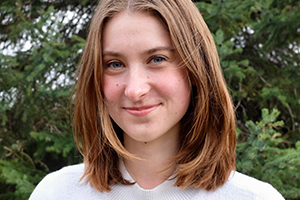 Clare Hermanson, with long brown hair, wearing a white shirt, with tree branches in the background.