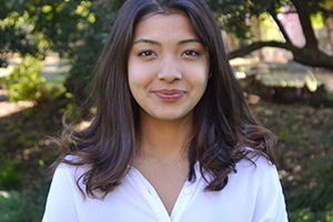 Nishtha Rajbhandari, with shoulder-length dark hair, wearing a white top, standing in the outdoors with trees in the background.