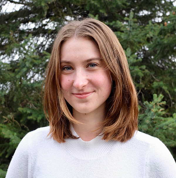 Clare Hermanson, with shoulder-length brown hair and wearing a white top, with foliage in the background.