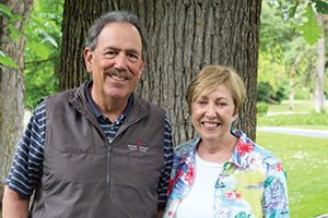 Roger and Rosemary Knapp stand next to each smiling. They are standing in front of a tree trunk and behind them is green grass and leaves.