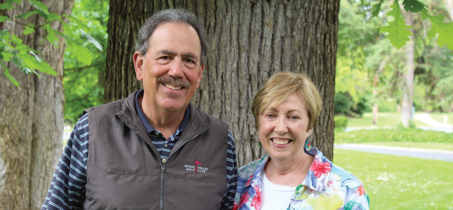 Roger and Rosemary Knapp stand next to each smiling. They are standing in front of a tree trunk and behind them is green grass and leaves.