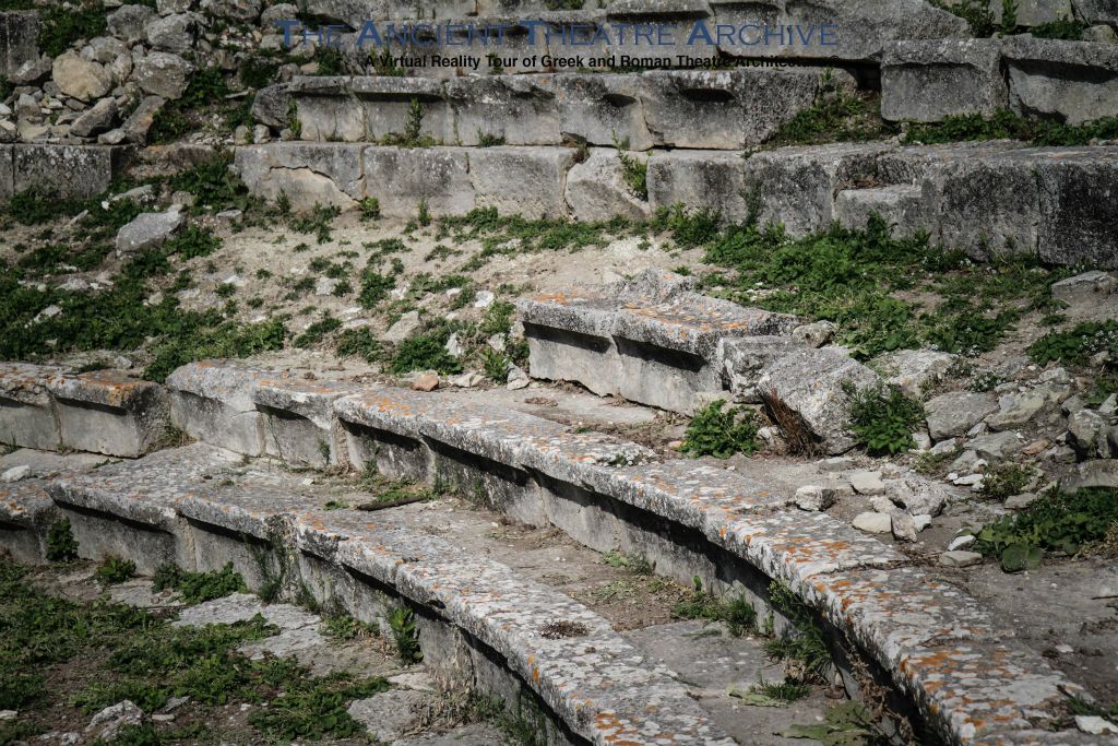 Orchestra: D 21.5 m, paved in beaten earth, surrounded by passageway (praecinctio) with 4 steps leading up from parodos; three rows of front seating (prohedriai), third row had continuous raised backs; both second and third rows have griffins’s foot terminations; reclining lion on low wall flanking ends of prohedriai; no drain around orchestra. Photo: T Hines 2019.